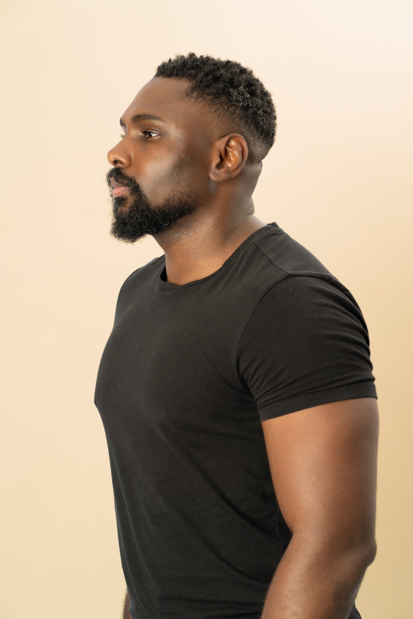 Chris Kibuka de profil, en t-shirt noir sur fond beige